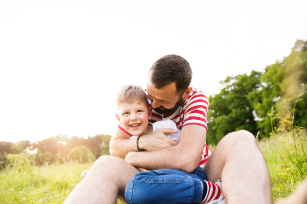 Young hipster father with his little son outside sitting on green meadow. Sunny summer day.