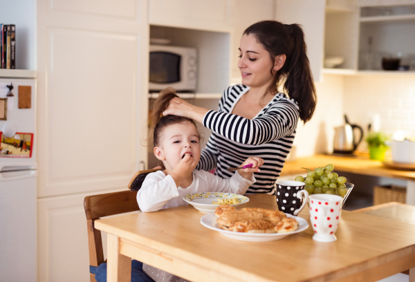 Cute little girl sitting at the table in the kitchen, eating breakfast. Mother doing her hair.