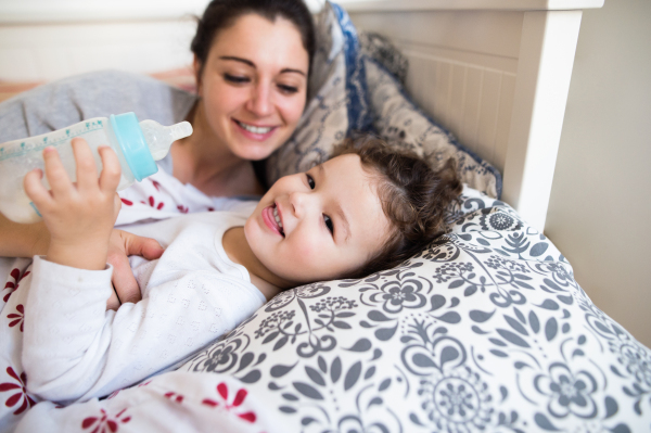 Beautiful young mother lying in bed, putting her cute little daughter to sleep, girl holding milk bottle.