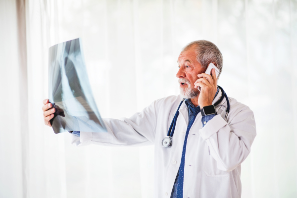 Senior doctor with smartphone looking at chest x-ray in his office. Male doctor examining an x-ray, talking on the phone.