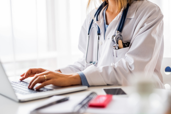 Unrecognizable young female doctor working on laptop at the office desk.