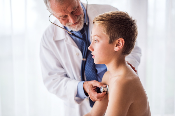 Senior male doctor examining a small boy with stethoscope in his office.