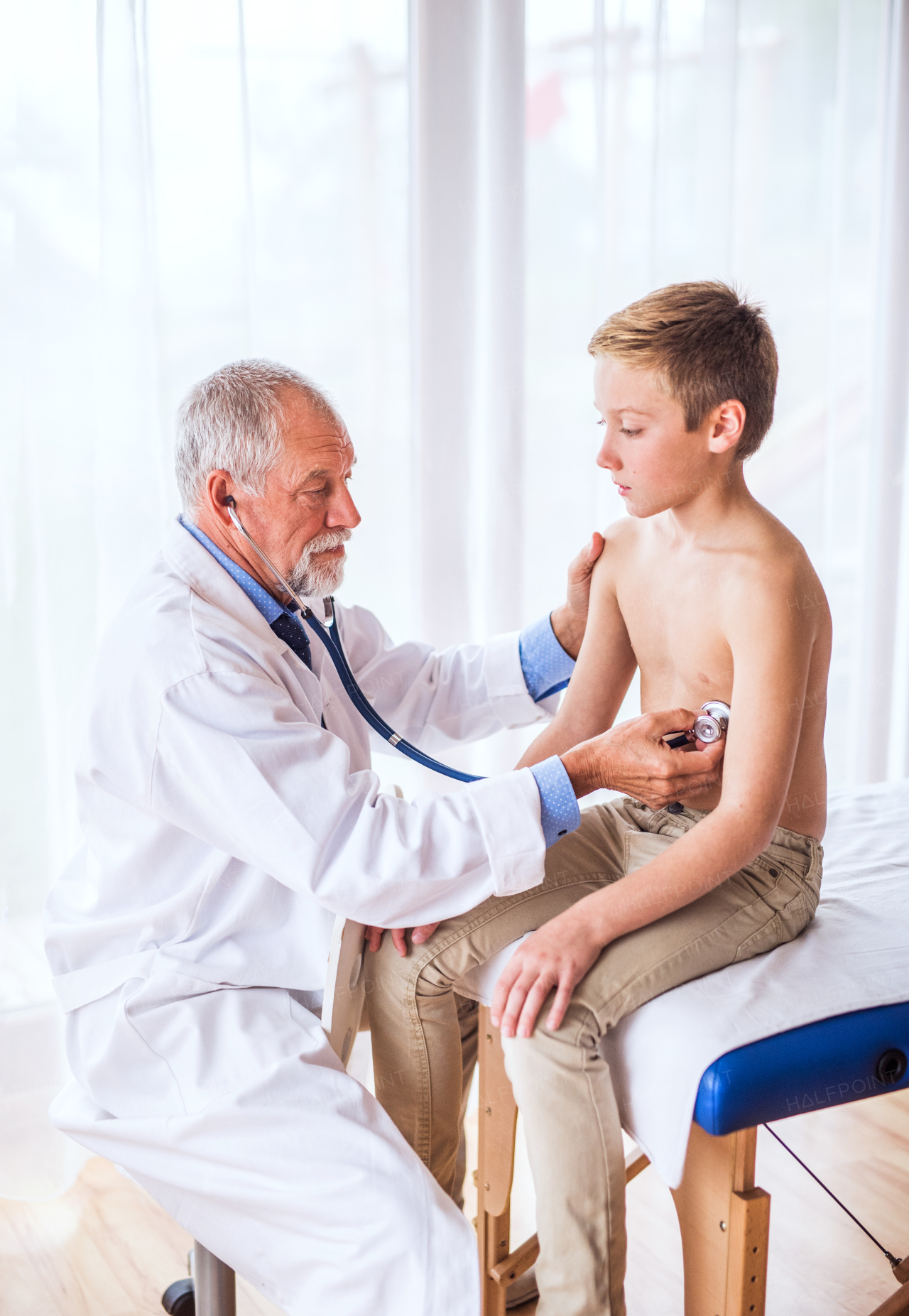 Senior male doctor examining a small boy with stethoscope in his office.