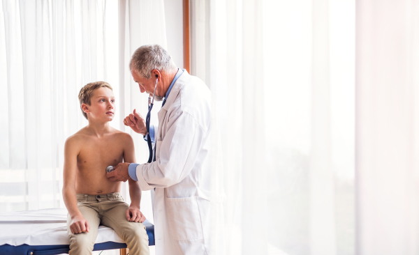 Senior male doctor examining a small boy with stethoscope in his office.