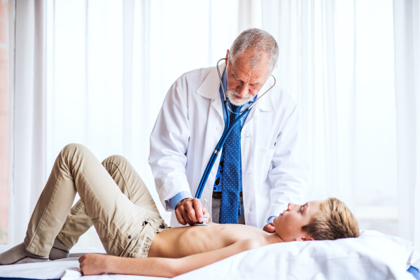 Senior male doctor examining a small boy with stethoscope in his office.