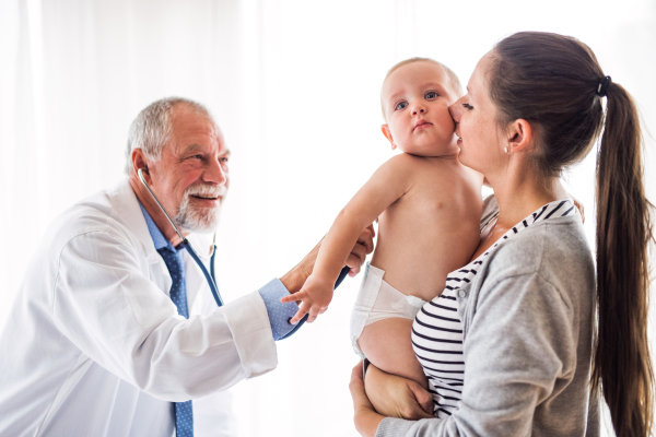 Senior male doctor examining a baby with stethoscope in his office. Young mother holding a boy.