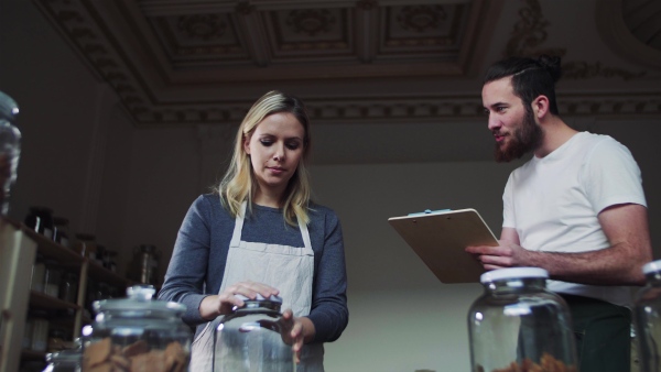 Two young male and female shop assistants with clipboard standing in zero waste shop, talking.