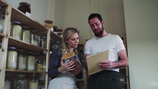 Two young male and female shop assistants with clipboard standing in zero waste shop, talking.