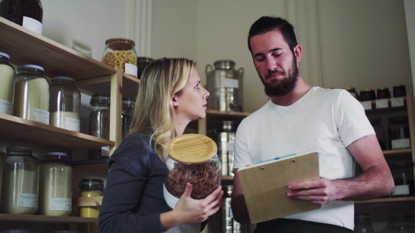 Two young male and female shop assistants with clipboard standing in zero waste shop, talking.