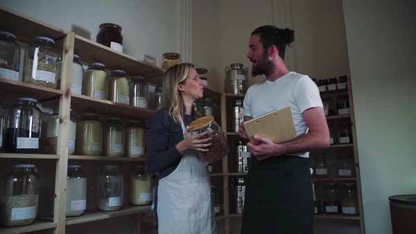 Two young male and female shop assistants with clipboard standing in zero waste shop, talking. Slow motion.