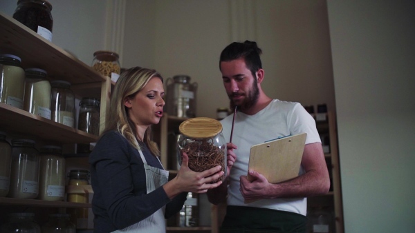 Two young male and female shop assistants with clipboard standing in zero waste shop, talking. Slow motion.