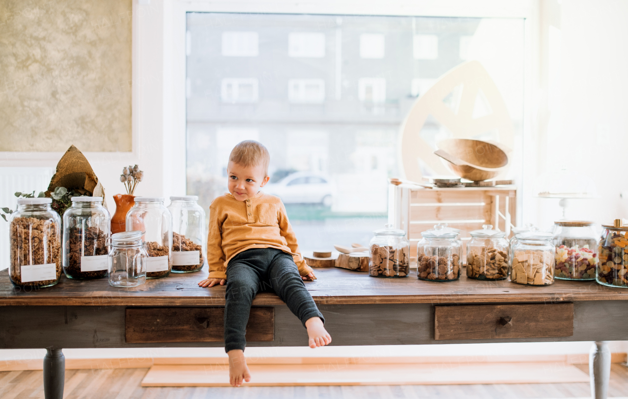 A happy barefoot small toddler boy sitting on a table in zero waste shop.