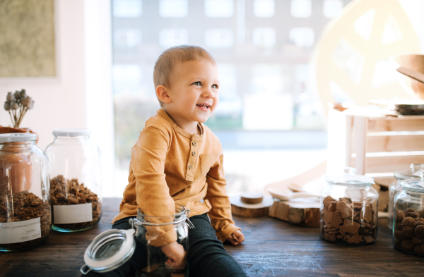 A happy small toddler boy sitting on a table in zero waste shop, holding a glass jar.