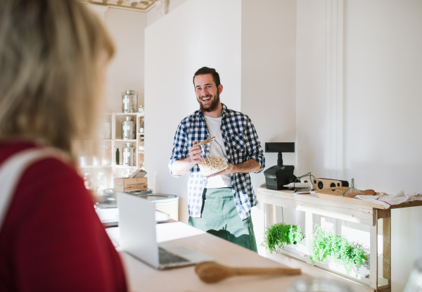 Young male shop assistant with laptop serving an unrecognizable woman in a zero waste shop.