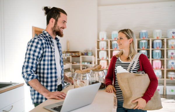 Young male shop assistant with laptop serving an attractive woman in a zero waste shop.