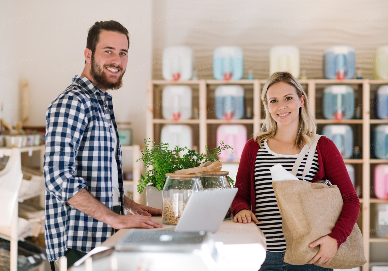 Young male shop assistant with laptop serving an attractive woman in a zero waste shop.