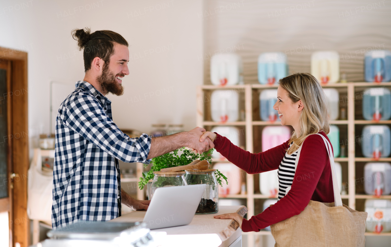 Young friendly male shop assistant with laptop and an attractive woman in a zero waste shop, shaking hands.