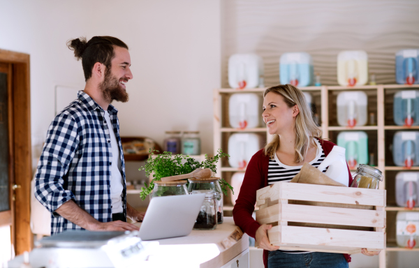 Young male shop assistant with laptop serving an attractive woman in a zero waste shop.