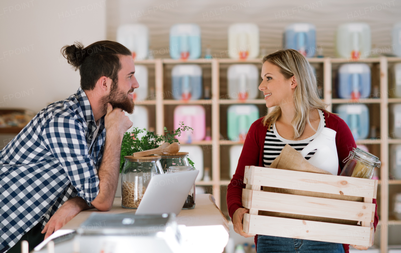 Young male shop assistant with laptop serving an attractive woman in a zero waste shop.