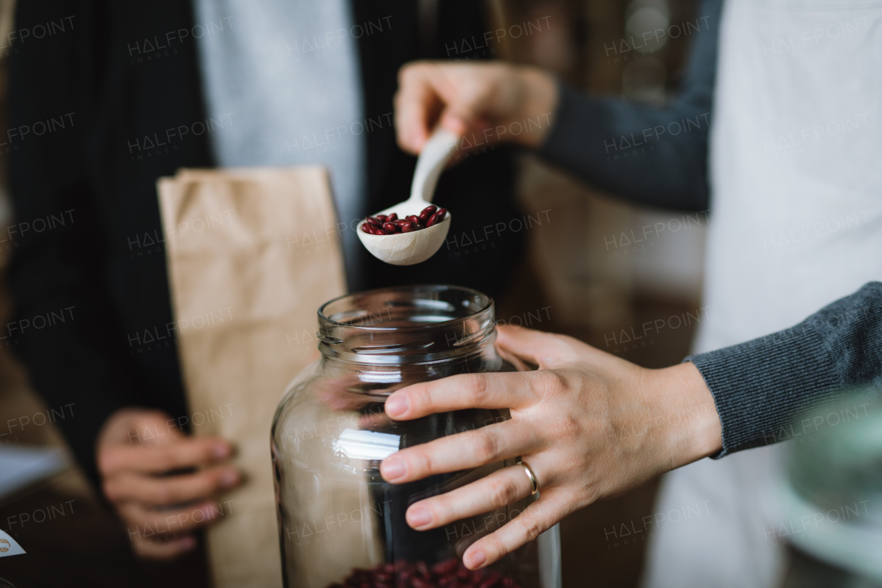 A midsection of a customer buying groceries in a zero waste shop, unrecognizable shop assitant putting beans in a paper bag. A close-up.