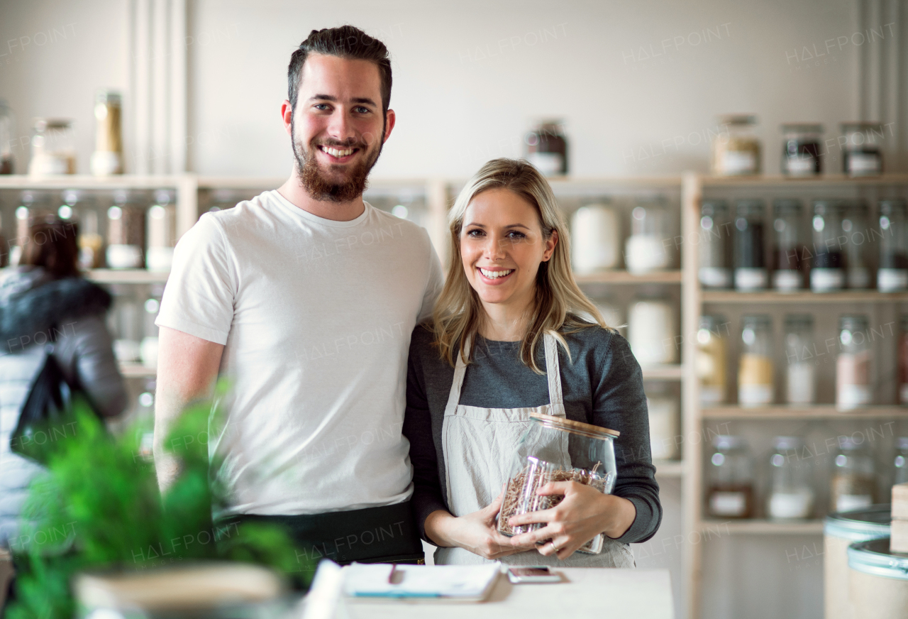 A portrait of two shop assistants standing arm in arm in zero waste shop, looking at camera.