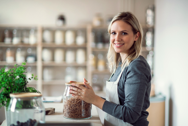 A young female shop assistant in a zero waste shop standing at the counter, holding a jar with groceries.