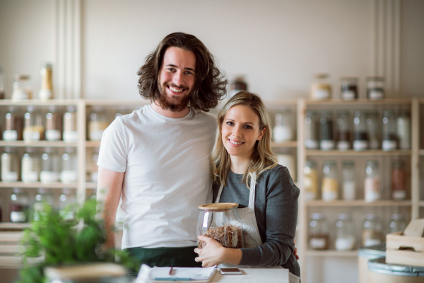 A portrait of two shop assistants standing arm in arm in zero waste shop, looking at camera.