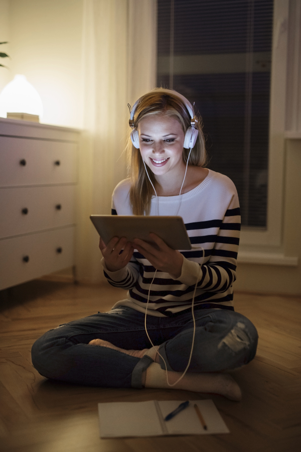 Young beautiful woman sitting on the floor, looking at the screen of a tablet late at night, headphones on head.