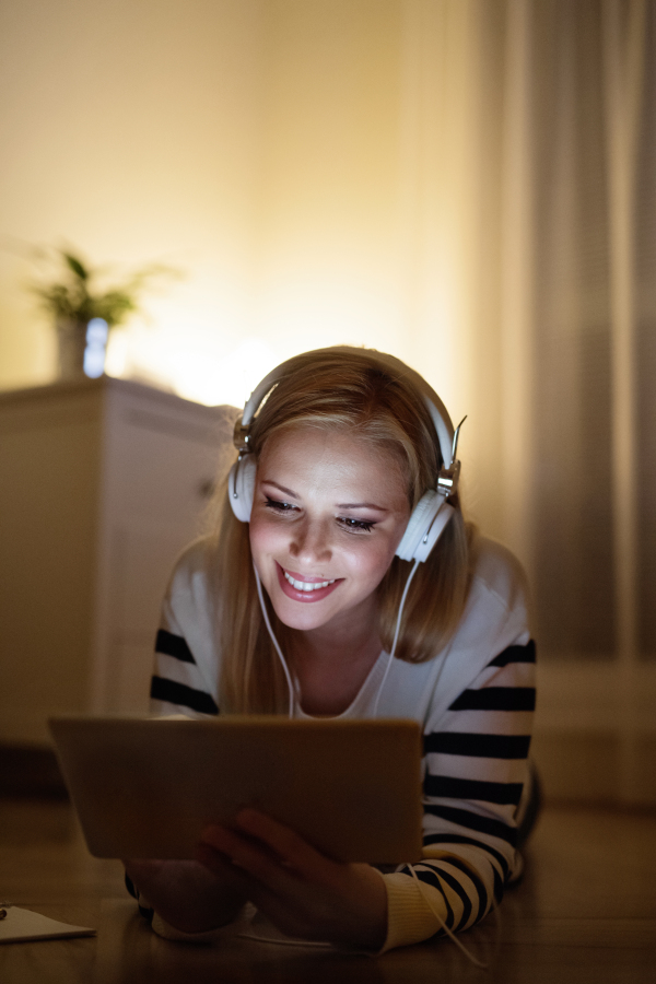 Young beautiful woman lying on the floor, looking at the screen of a tablet late at night, headphones on head.