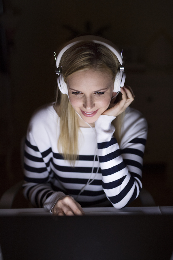 Young beautiful woman in striped sweater sitting at desk, looking at the screen of a laptop late at night, headphones on head.