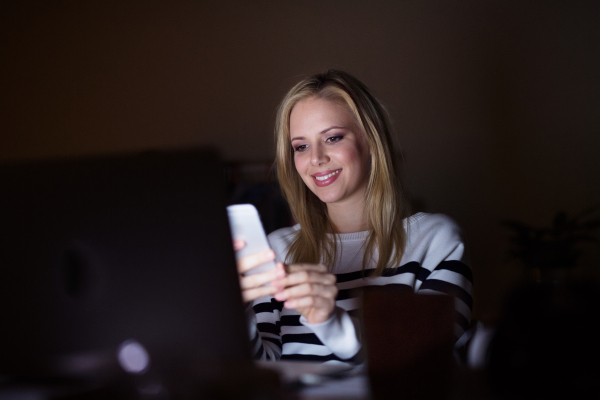 Young beautiful woman sitting at desk at night, laptop in front of her, holding smart phone, texting.