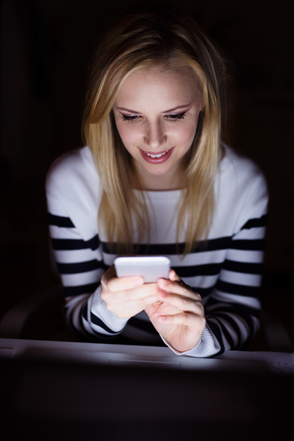 Young beautiful woman sitting at desk at night, laptop in front of her, holding smart phone, texting.