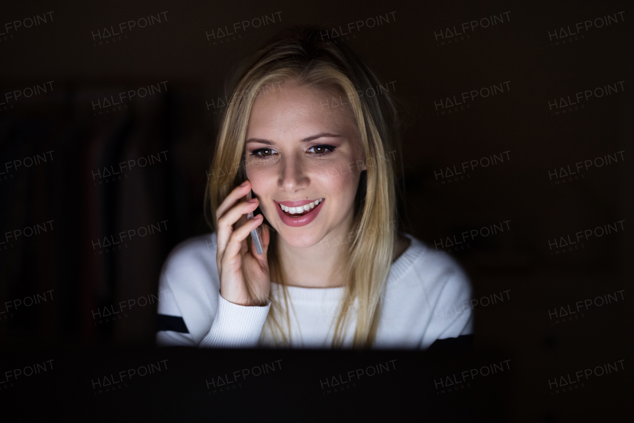 Beautiful young blonde woman sitting at desk at night, holding smart phone, making phone call.