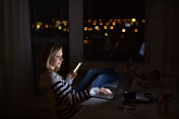 Young beautiful woman sitting at desk at night, holding smartphone, working on laptop at night.