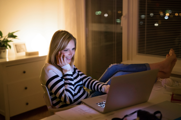 Young beautiful woman sitting at desk at night, holding smartphone, making phone call, working on laptop.