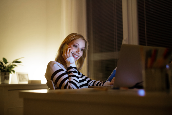 Young beautiful woman sitting at desk at night, laptop in front of her, holding smart phone, making phone call