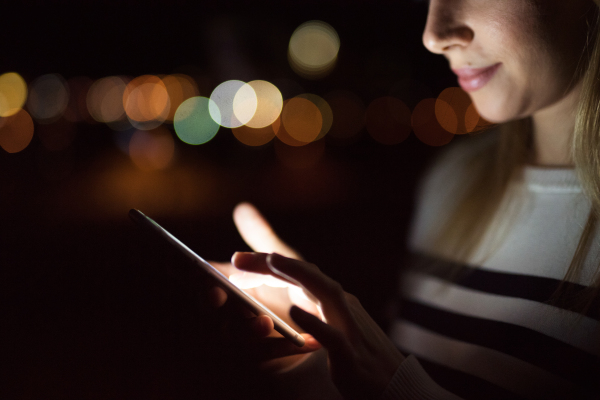 Unrecognizable young woman sitting at desk at night, holding smart phone, texting.