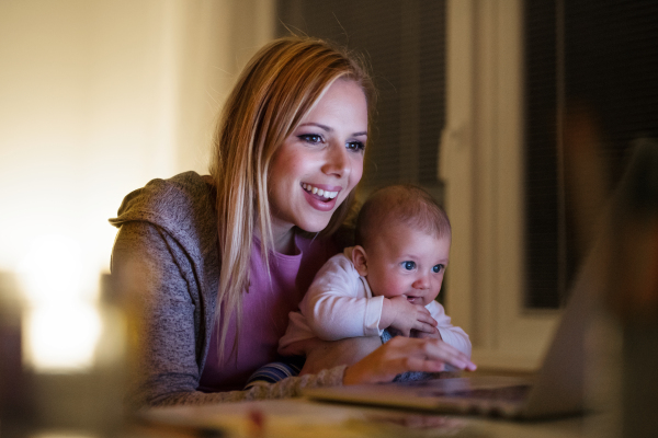 Beautiful young mother at home at night with her little baby son in the arms, working on laptop