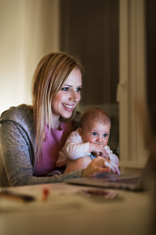 Beautiful young mother at home at night with her little baby son in the arms, working on laptop