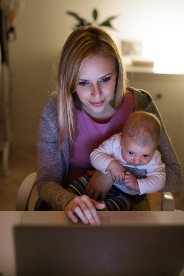 Beautiful young mother at home at night with her little baby son in the arms, working on laptop