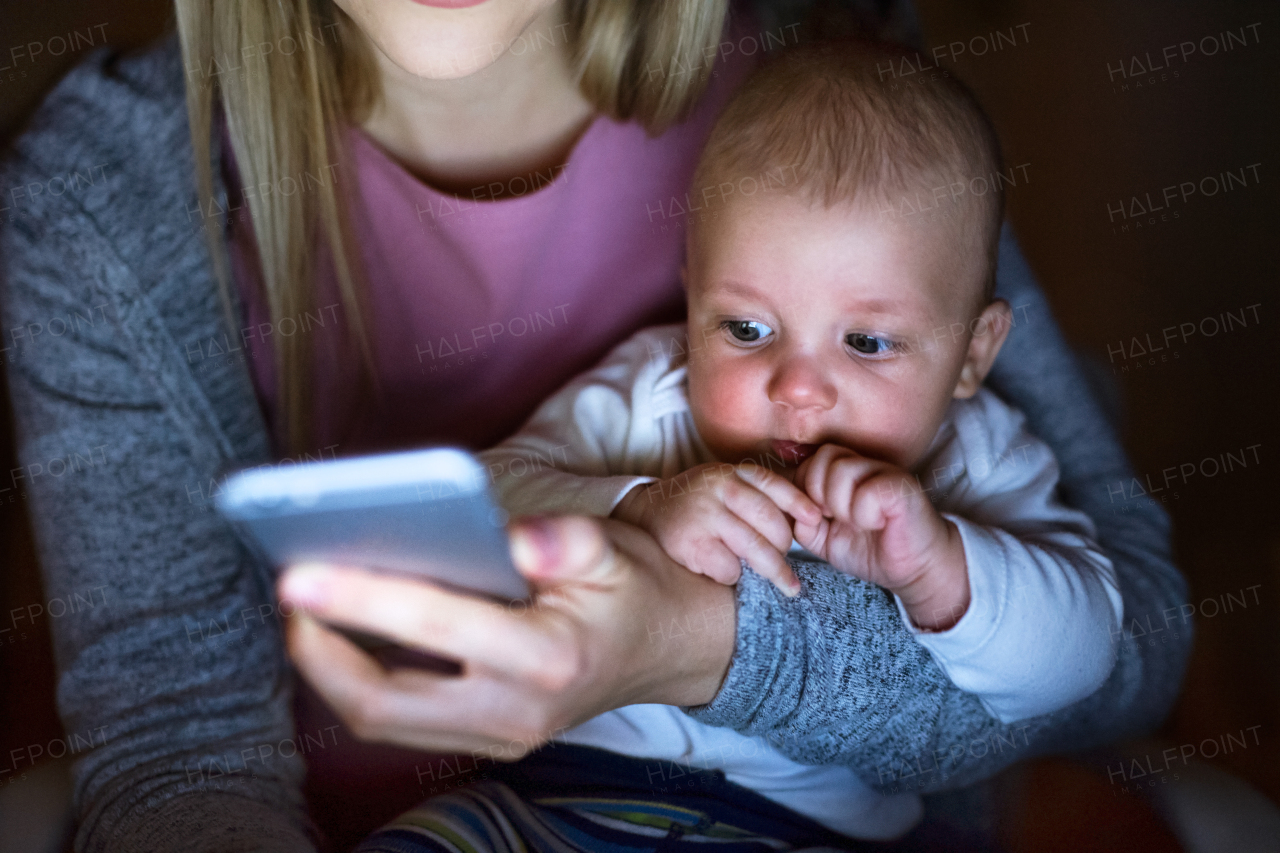 Unrecognizable young blond mother at home at night with her little baby son in the arms, holding smart phone, texting