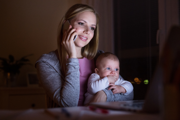 Beautiful young mother at home at night with her little baby son in the arms, sitting in front of laptop, holding smart phone, making phone call