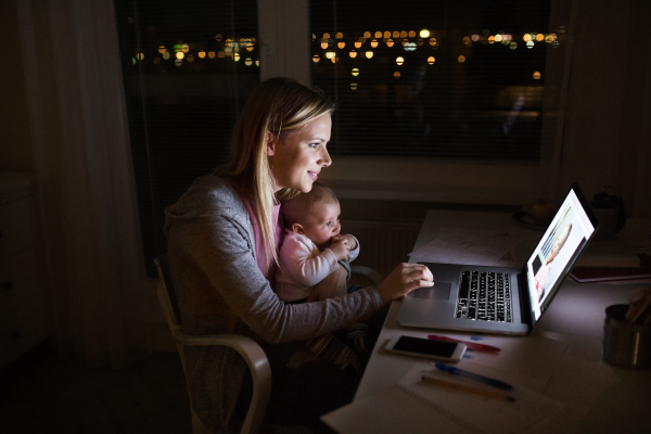 Beautiful young mother at home at night with her little baby son in the arms, working on laptop