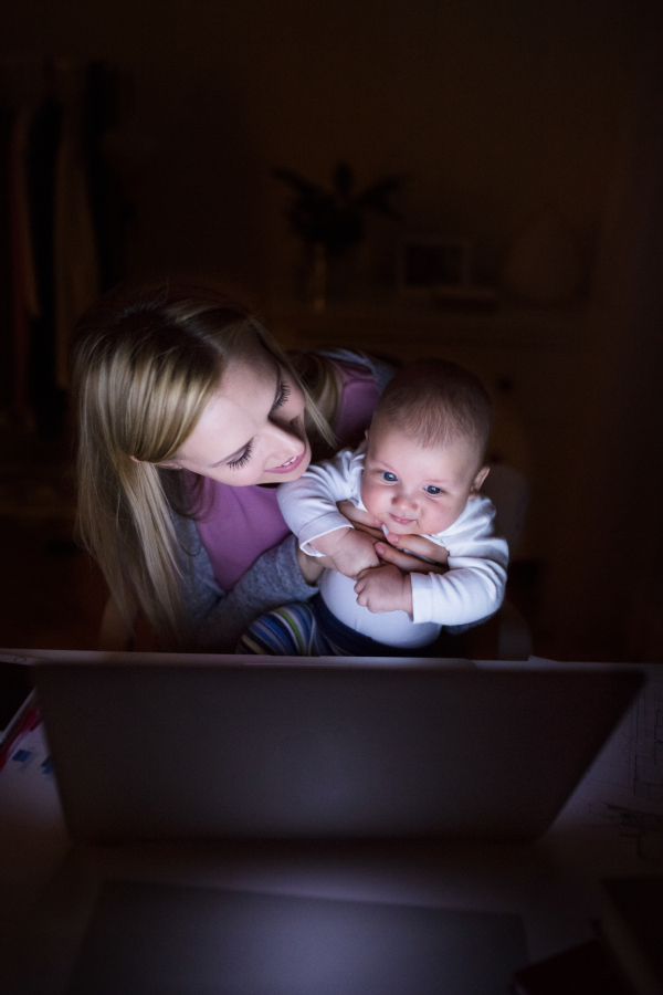 Beautiful young mother at home at night with her little baby son in the arms, working on laptop