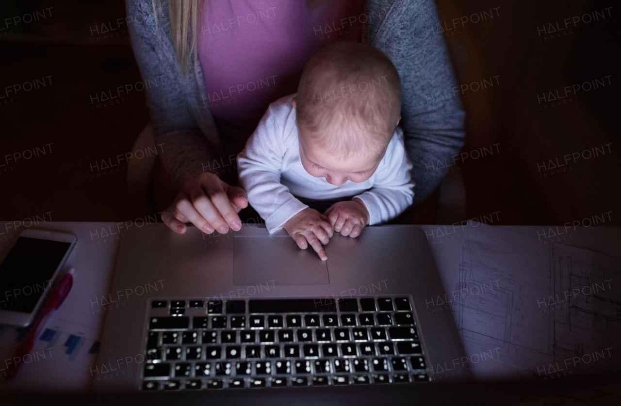 Unrecognizable young mother at home at night with her little baby son sitting on her lap, working on laptop