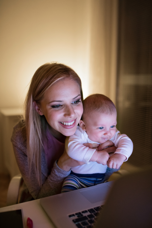 Beautiful young mother at home at night with her little baby son in the arms, working on laptop