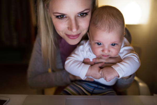 Beautiful young mother at home at night with her little baby son in the arms, laptop in front of her