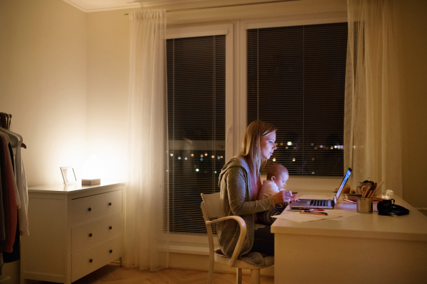 Beautiful young mother at home at night with her little baby son in the arms, working on laptop