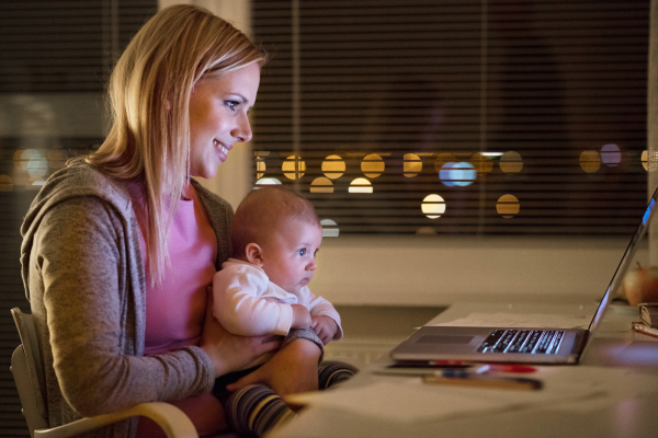 Beautiful young mother at home at night with her little baby son in the arms, working on laptop, smiling