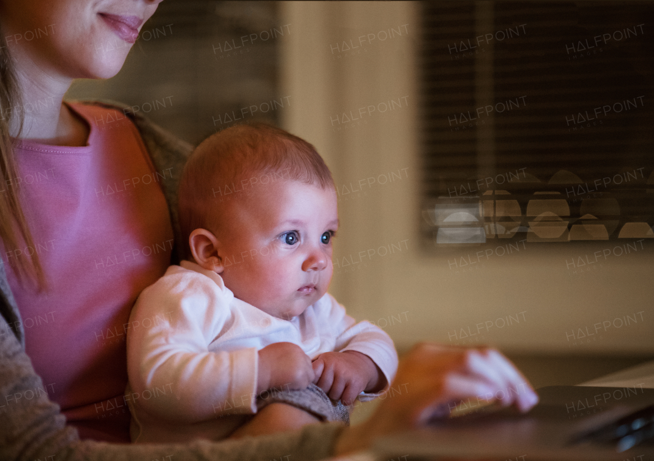 Unrecognizable young mother at home at night with her little baby son sitting on her lap, working on laptop